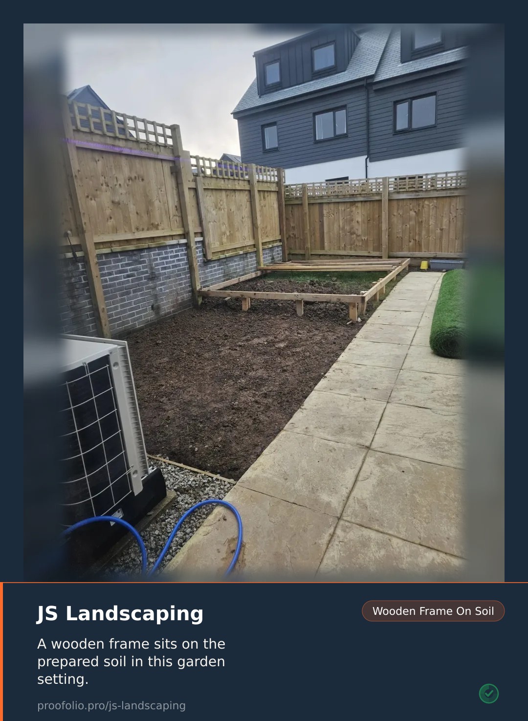 A wooden subframe sits on a patch of cleared earth next to a stone patio and a roll of artificial grass.
